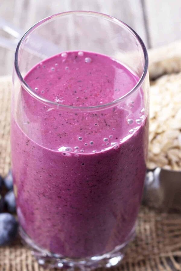 Cholesterol-Lowering Blueberry Oat smoothie in a glass, on my kitchen counter, surrounded by fresh fruit and vegetables.