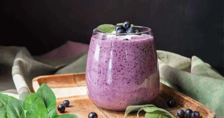 Cholesterol-Lowering Blueberry Spinach smoothie in a glass, on my kitchen counter, surrounded by fresh fruit and vegetables.