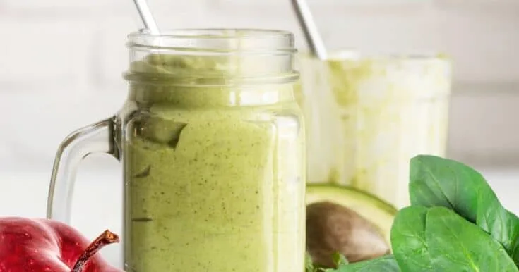 Cholesterol-Lowering Green Avocado smoothie in a glass, on my kitchen counter, surrounded by fresh fruit and vegetables.