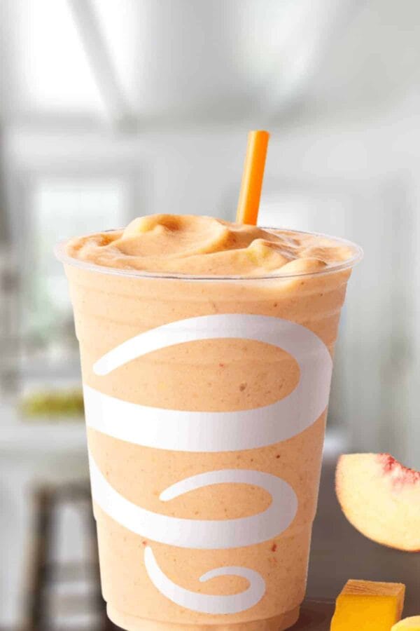Jamba Juice Peach Mango smoothie in a glass, on my kitchen counter, surrounded by fresh peaches and mango chunks.