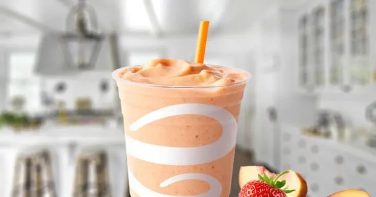 Jamba Juice Peach Perfection smoothie in a glass, on my kitchen counter, surrounded by fresh peaches and berries.