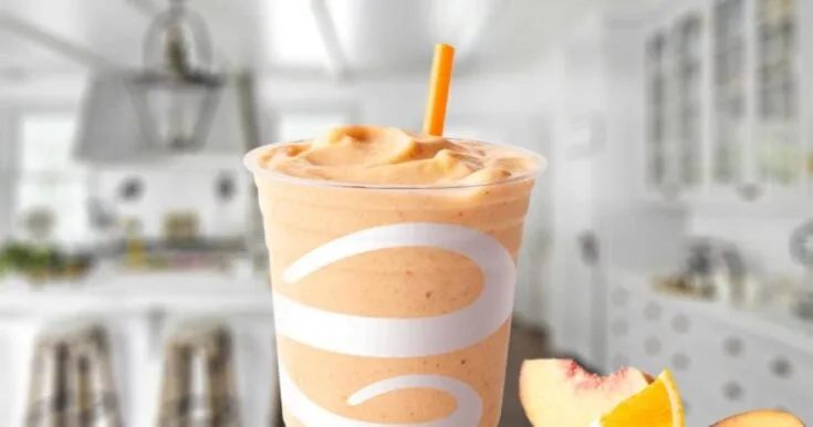 Jamba Juice Peach Pleasure smoothie in a glass, on my kitchen counter, surrounded by fresh peaches and orange slices.