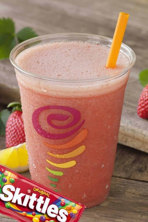 Jamba Juice Secret Menu Skittles smoothie in a glass, on my backyard table, surrounded by fresh fruit and skittles candy.