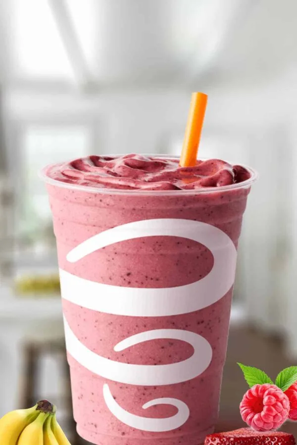 Jamba Juice Strawberry Raspberry Banana smoothie in a glass, on my kitchen counter, surrounded by fresh berries and banana slices.