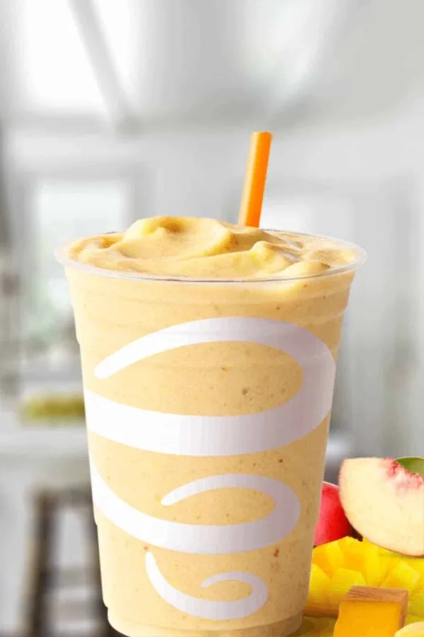 Jamba Juice Tropical Harvest smoothie in a glass, on my kitchen counter, surrounded by fresh peaches and mango.