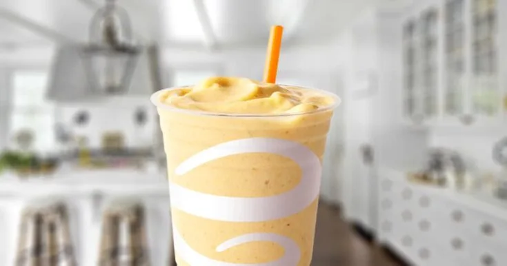 Jamba Juice Tropical Harvest smoothie in a glass, on my kitchen counter, surrounded by fresh peaches and mango.