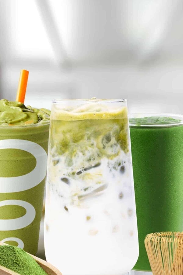 Matcha smoothies in glasses, on my kitchen counter, surrounded by green tea matcha powder and a bamboo whisk.