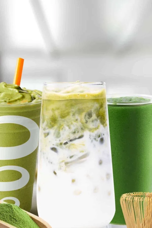 Matcha smoothies in glasses, on my kitchen counter, surrounded by green tea matcha powder and a bamboo whisk.