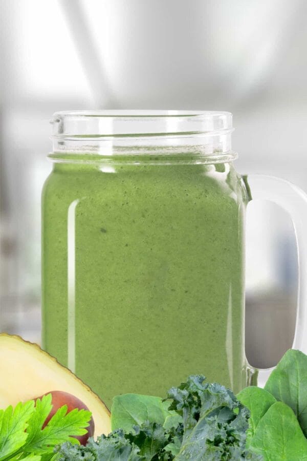 Green smoothie in a glass, on my kitchen counter, surrounded by fresh vegetables.