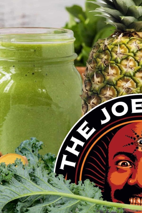 Joe Rogan Kale Shake smoothie in a glass, on my kitchen counter, surrounded by fresh fruit, vegetables, and a Joe Rogan Podcast sticker.