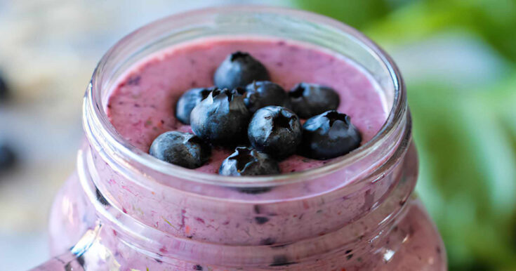 NutriBullet Blueberry Forever smoothie in a glass, on my kitchen counter, surrounded by fresh fruit and vegetables.