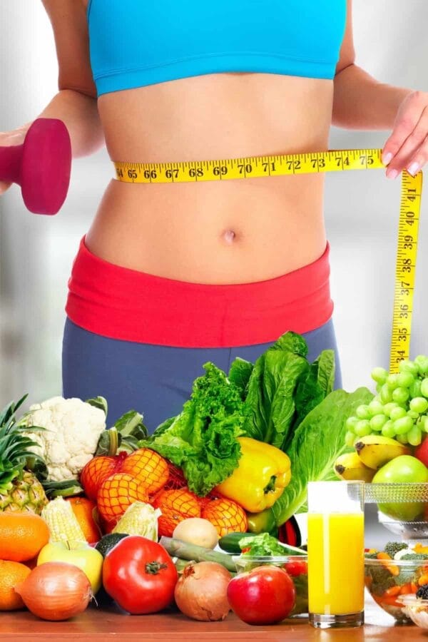 Fit woman with a flat belly, standing next to a weight loss smoothie, surrounded by fresh fruit and vegetables.