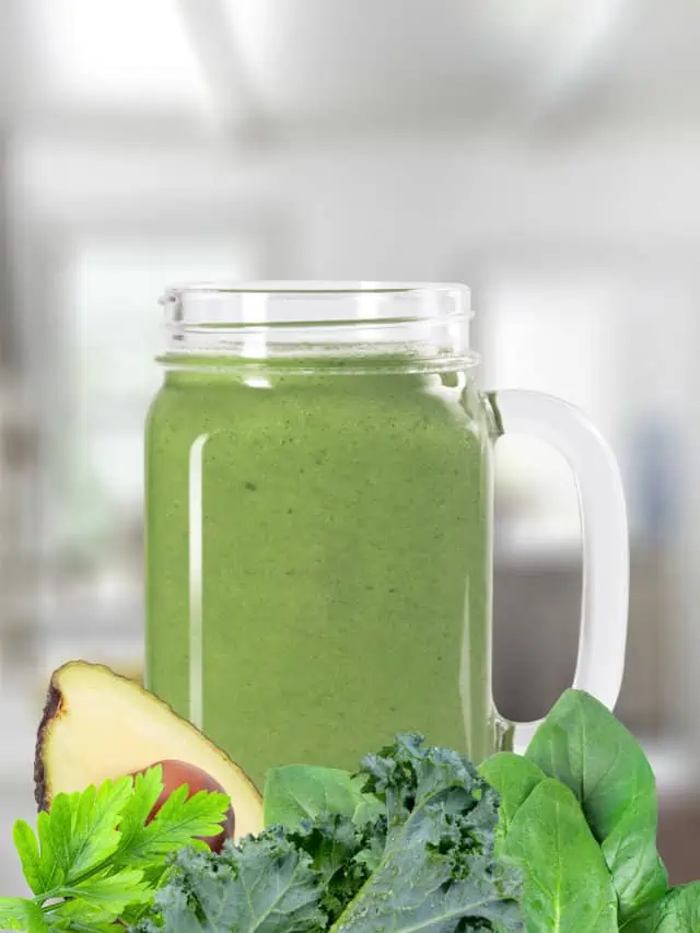 Green smoothie in a glass, on my kitchen counter, surrounded by fresh vegetables.