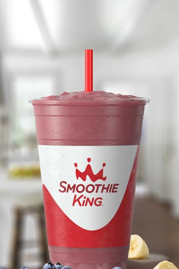 Smoothie King Blueberry Heaven smoothie in a glass, on my kitchen counter, surrounded by fresh bananas and blueberries.