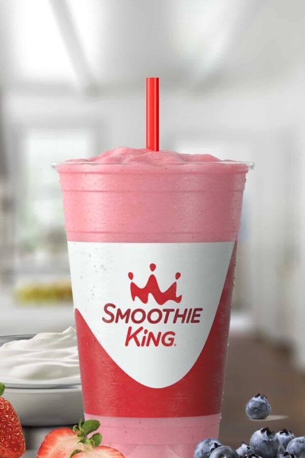 Smoothie King Kids Strawberry Bluegurt Blitz smoothie in a glass, on my kitchen counter, surrounded by fresh berries and Greek yogurt.