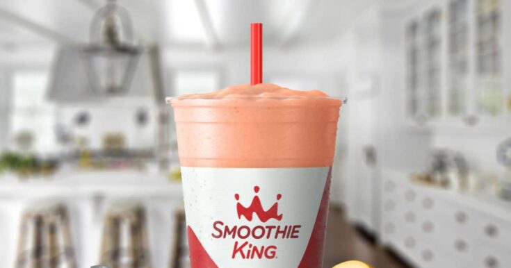 Smoothie King Kindness In A Cup smoothie in a glass, on my kitchen counter, surrounded by fresh fruit and whey protein powder.