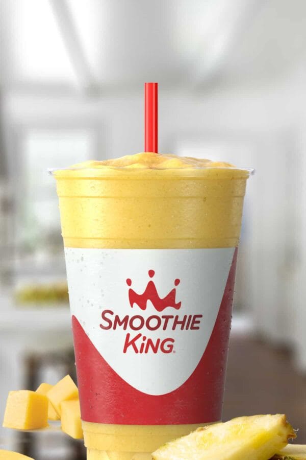 Smoothie King MangoFest smoothie in a glass, on my kitchen counter, surrounded by fresh mango and pineapple slices.