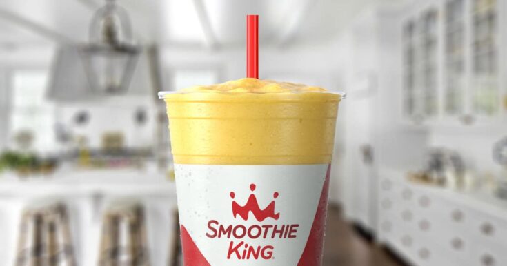 Smoothie King MangoFest smoothie in a glass, on my kitchen counter, surrounded by fresh mango and pineapple slices.