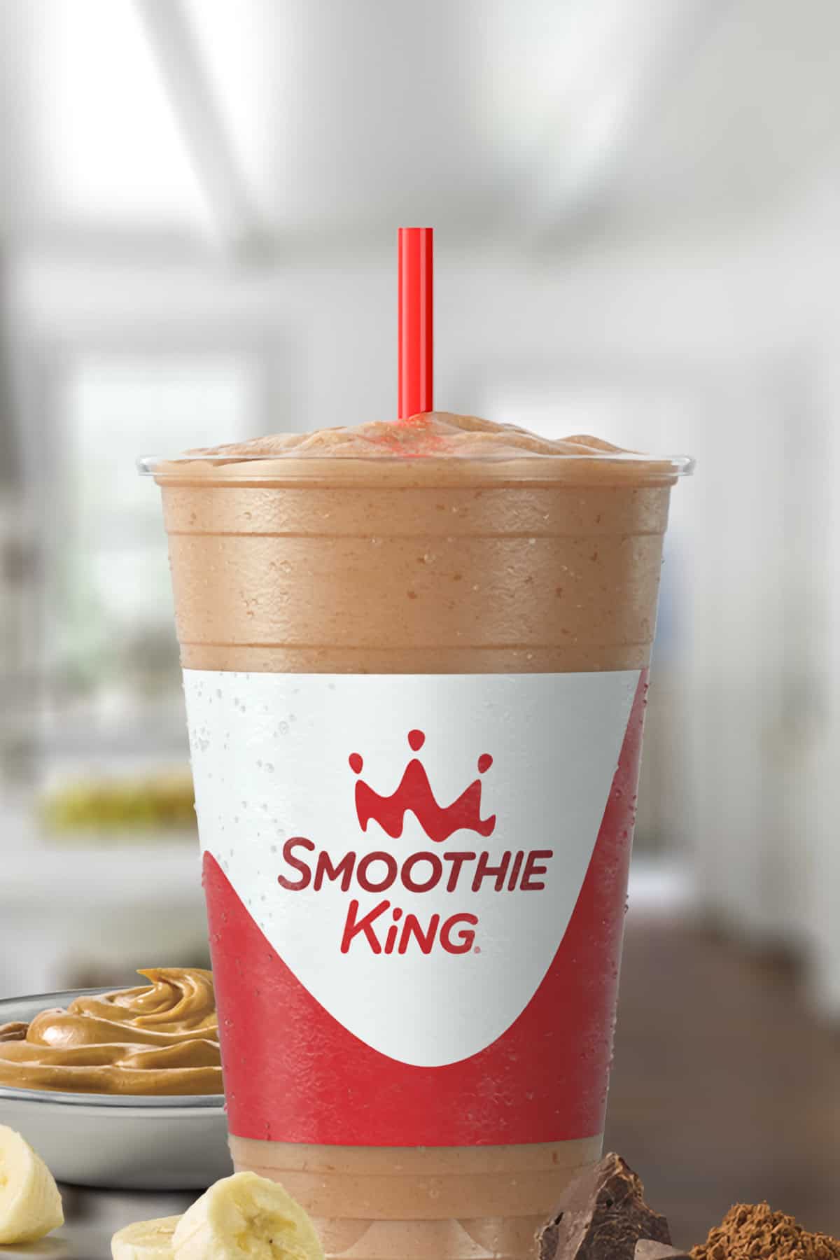 Smoothie King Peanut Power Plus Chocolate smoothie in a glass, on my kitchen counter, surrounded by creamy peanut butter, sliced bananas, and cacao.