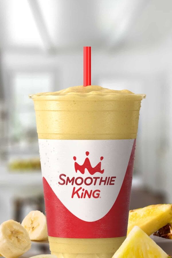 Smoothie King Pure Recharge Pineapple smoothie in a glass, on my kitchen counter, surrounded by fresh bananas and pineapples.