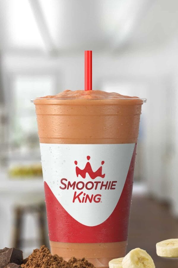 Smoothie King The Activator Recovery Chocolate smoothie in a glass, on my kitchen counter, surrounded by cacao powder and sliced bananas.